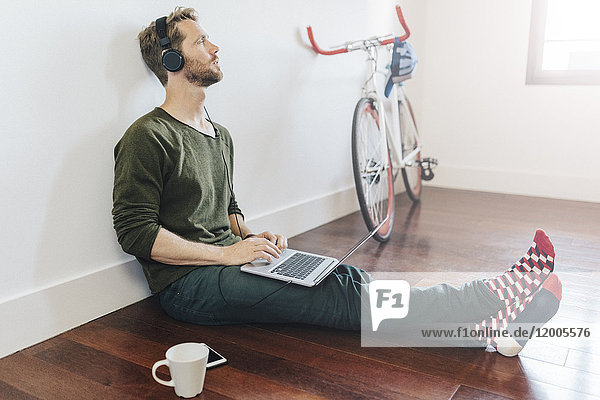 Nachdenklicher Mann mit Kopfhörer und Laptop zu Hause auf dem Boden sitzend