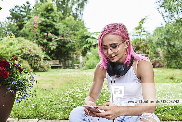 Junge Frau mit rosa Haaren mit Kopfhörer und Handy im Garten