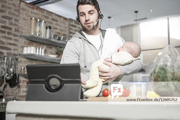 Father with headset and tablet in kitchen holding baby