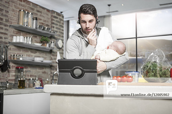 Vater mit Kopfhörer und Tablette in der Küche hält Baby