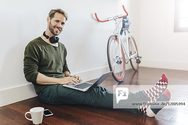 Porträt eines lächelnden Mannes mit Kopfhörer zu Hause auf dem Boden sitzend mit Laptop