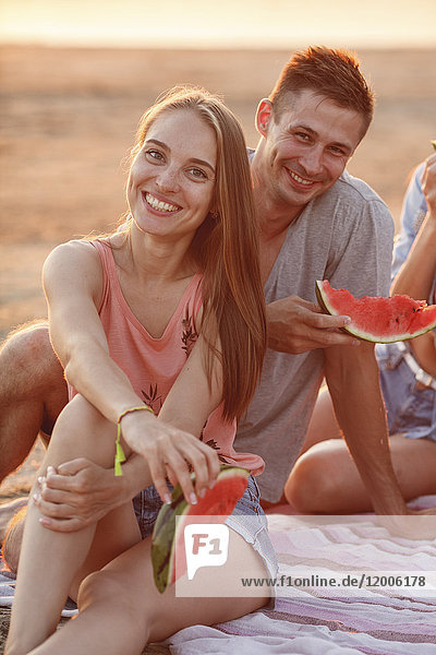 Glückliches Paar mit Freunden am Strand beim Essen von Wassermelone