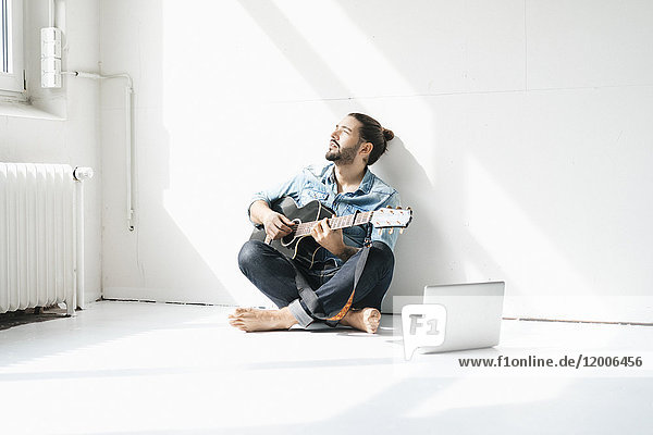 Mann sitzt mit Laptop auf dem Boden in einem Loft und spielt Gitarre.