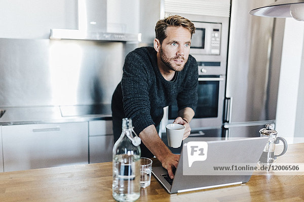 Portrait des Hauptdarstellers mit Kaffeetasse und Laptop in der Küche stehend