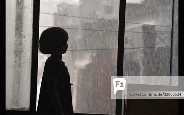 Silhouette eines jungen Mädchens,  das durch ein Fenster den Regen beobachtet