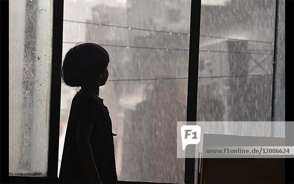 Silhouette eines jungen Mädchens  das durch ein Fenster den Regen beobachtet