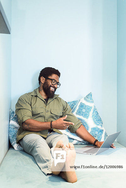 Lächelnder schwarzer Mann sitzt auf einem Bett und zeigt auf einen Laptop