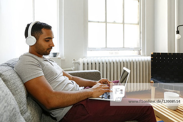Gemischtrassiger Mann sitzt auf dem Sofa und hört mit Kopfhörern auf seinen Laptop
