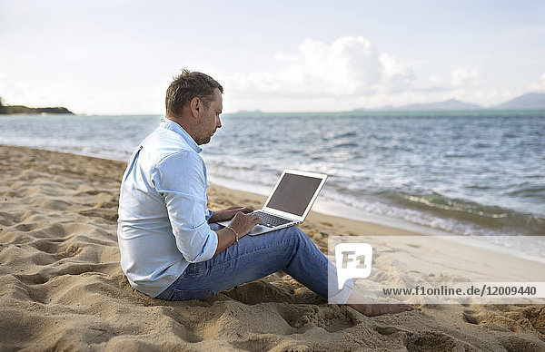 Kaukasischer Mann sitzt am Strand und benutzt einen Laptop
