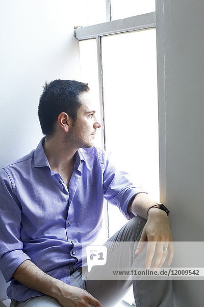 Nachdenklicher kaukasischer Mann sitzt auf der Fensterbank