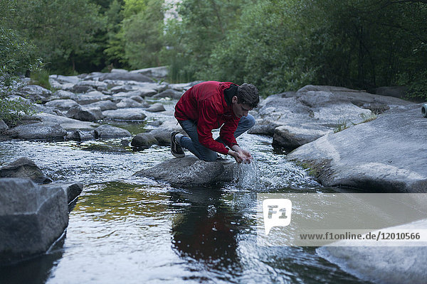 Kaukasischer Mann kniet auf einem Felsen im Fluss und schröpft Wasser mit den Händen