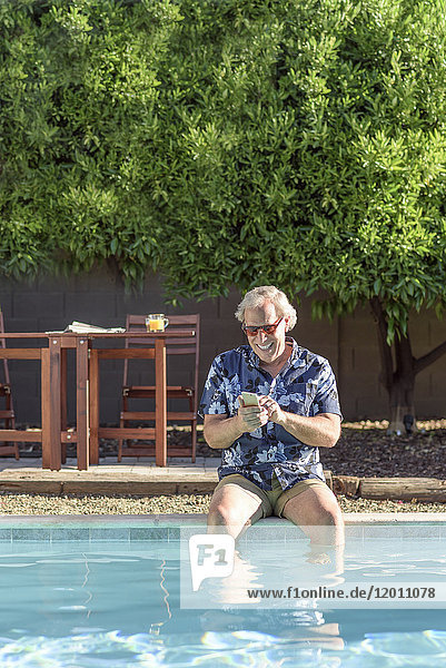 Caucasian man texting on cell phone with legs in swimming pool