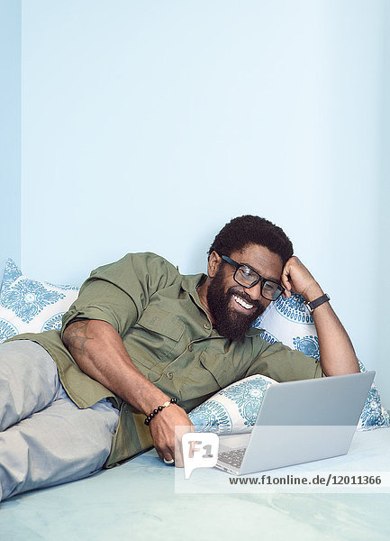 Lächelnder schwarzer Mann auf dem Bett liegend mit Laptop