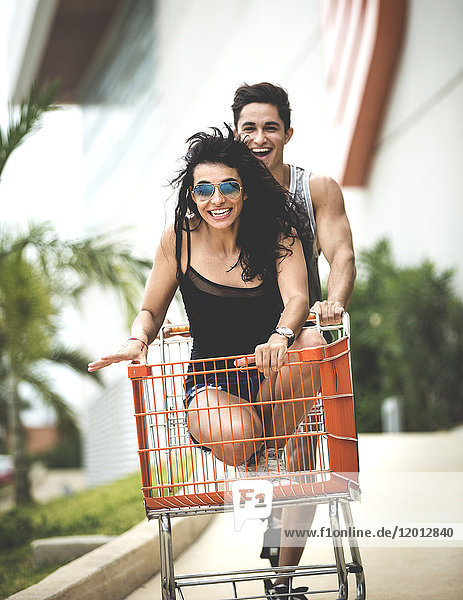 A young man pushing a young woman sitting in a shopping trolley.