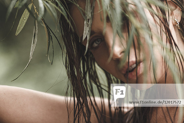 Nahaufnahme einer jungen Frau mit nassen Haaren  die durch die Blätter schaut.