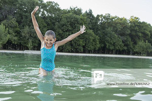 Fröhliches Mädchen mit erhobenen Armen  das im Schwimmbad gegen Bäume genießt.