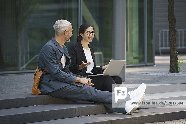 Smiling business colleagues with laptop discussing while sitting on steps in city