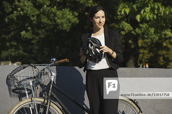 Lächelnde Geschäftsfrau  die an einem sonnigen Tag den Helm am Fahrrad gegen die Pflanzen hält.
