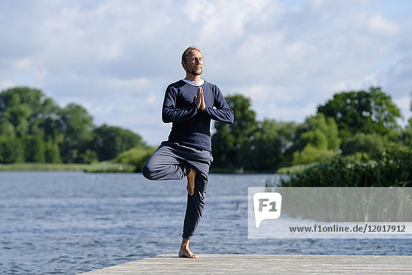 Reifer Mann praktiziert Baum-Pose Yoga auf Pier am See gegen den Himmel