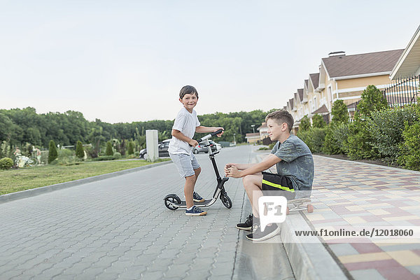 Junge sitzt auf dem Skateboard  während sein Bruder auf der Kopfsteinpflasterstraße Roller fährt.