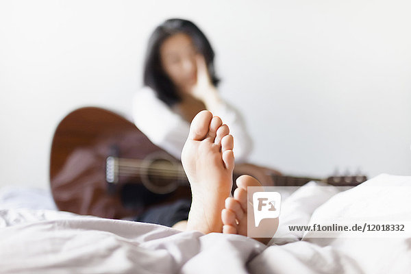 Frau mit nackten Füßen hält die Gitarre  während sie zu Hause auf dem Bett liegt.