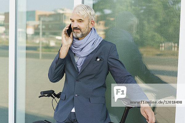 Confident businessman with bicycle talking on mobile phone while standing against glass building