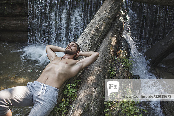 High angle view of thoughtful shirtless man lying on logs over waterfall at forest