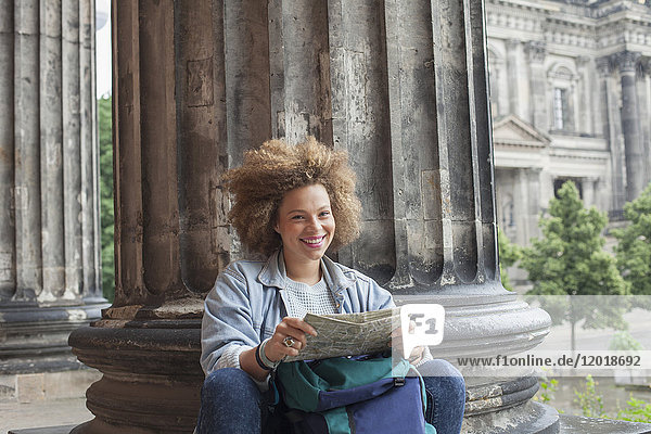 Portrait of smiling young Afro female tourist sitting with map against column at Altes Museum