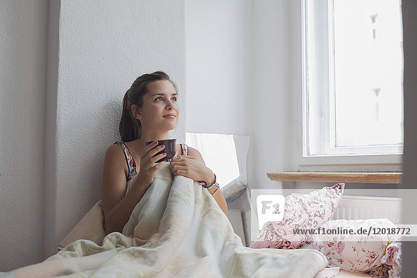 Nachdenkliche junge Frau sitzend mit Kaffeetasse auf dem Bett zu Hause