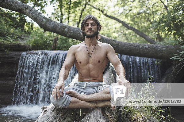 Der Mann ohne Hemd sitzt mit gekreuzten Beinen auf Baumstämmen gegen den Wasserfall im Wald.