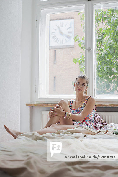 Schöne junge Frau sitzt zu Hause auf dem Bett gegen das Fenster.