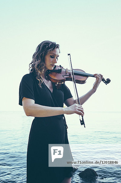 Schöne junge Frau spielt Geige  während sie am See gegen den klaren Himmel steht.