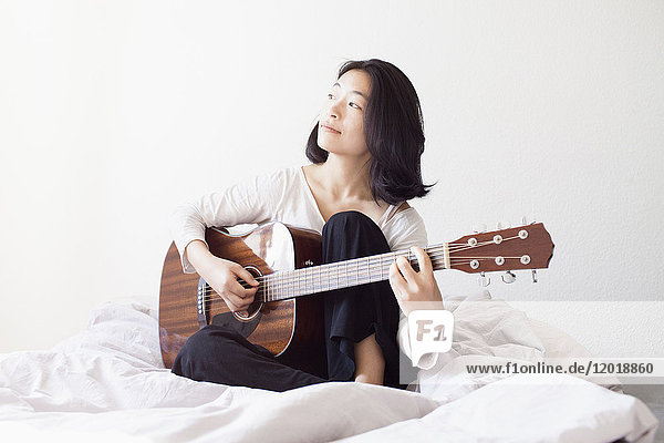 Nachdenkliche Frau spielt Gitarre  während sie zu Hause auf dem Bett sitzt.