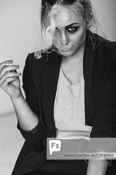 Portrait of young woman smoking cigarette while sitting against gray background