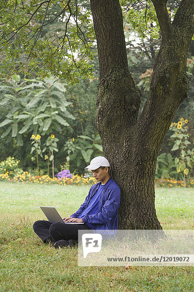 Volle Länge des Mannes  der den Laptop benutzt  während er am Baum auf einer Wiese im Park sitzt.