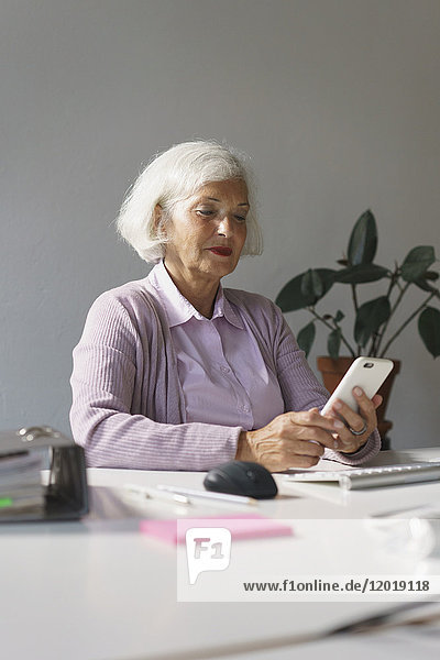 Smiling senior woman looking at mobile phone while sitting against wall in office