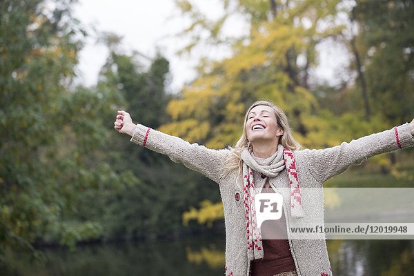 Porträt einer jungen blonden Frau  die an einem See im Herbst mit den Armen in die Luft lächelt.