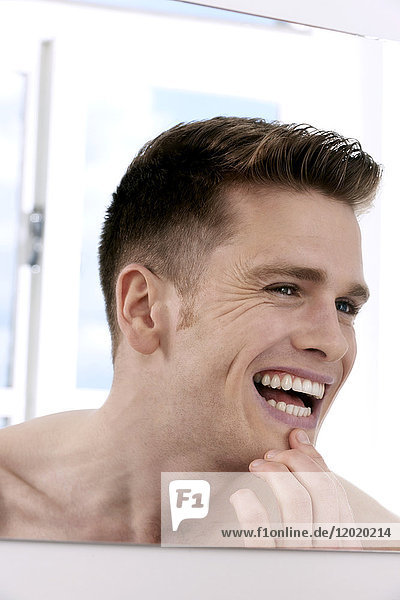Oben-ohne-Mann  schaut in den Spiegel  betrachtet seine Zähne im Spiegel  Finger am Kinn  lächelnd  Fenster im Hintergrund