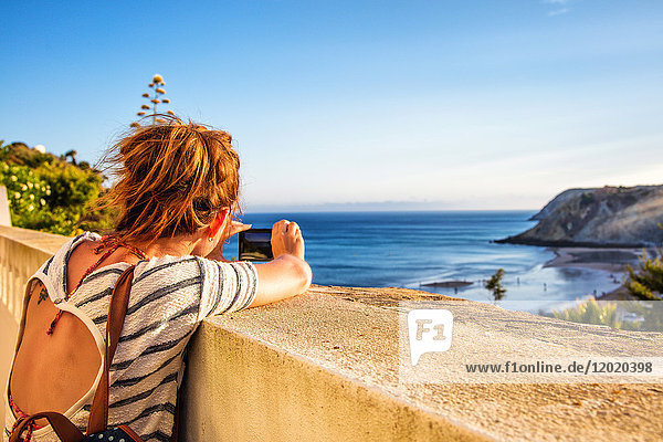 Junge Frau von hinten  nimmt ein Foto mit seinem Smartphone auf einer niedrigen Mauer vom Strand von Burgau  Region Algarve  Portugal