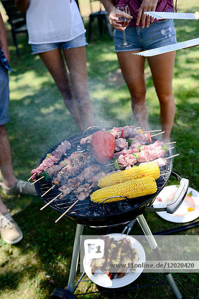 Close up of a barbecue grill with meat and sausages cooking during a summer garden party with people in background.