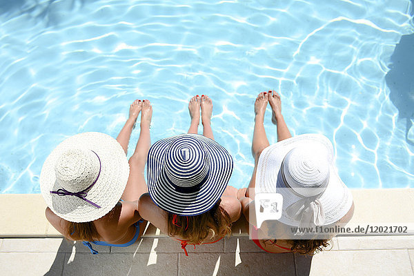 Drei schöne junge Frau mit Badeanzug und Sonnenhut sitzen am Rande des Pools der Anlage Schwimmbad während des Sommerurlaubs.