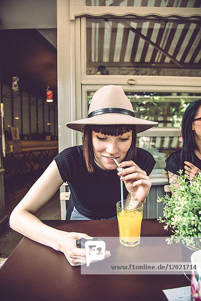 Porträt einer jungen Frau  die auf einer Restaurantterrasse sitzt  ein Mobiltelefon berät und Fruchtsaft trinkt