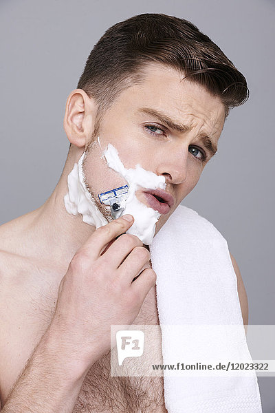 Oben-ohne-Mann  Handtuch auf der Schulter  mit Rasierschaum auf der Gesichtshälfte  mit Rasiermesser in den Händen  ernst  rasierend  offener Mund