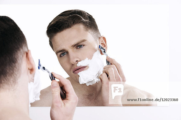 Oben-ohne-Mann  in den Spiegel schauend  rasierend  mit Rasierapparat in der Hand  Rasierschaum auf der Hälfte seines Gesichts