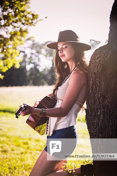 Junge Frau auf dem Rücken eines Baumes spielt Gitarre
