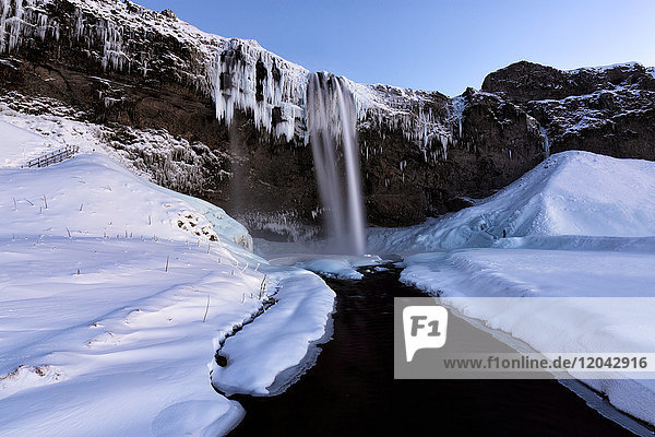Winter view of Seljalandsfoss Waterfall at dusk with snow covered foreground and icicles hanging from cliffs South Iceland Polar Regions