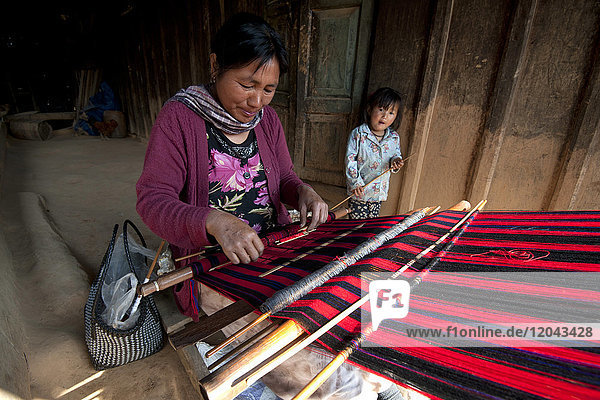 Frau beim Weben des traditionellen roten Naga-Schals  daneben ihre kleine Tochter  auf der Veranda des Familienhauses  Nagaland  Indien  Asien