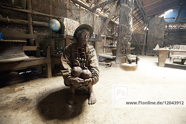 Naga man Longsha Wangnao head hunter with tattooed face and Naga necklace sitting in communal Naga tribal house Longwa Nagaland India Asia
