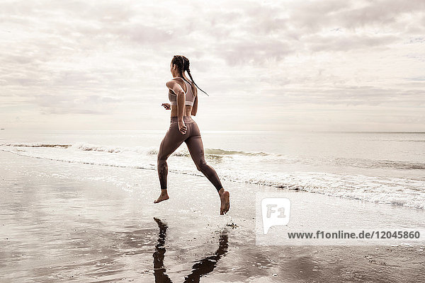 Rear view of young female runner running barefoot along water's edge at beach