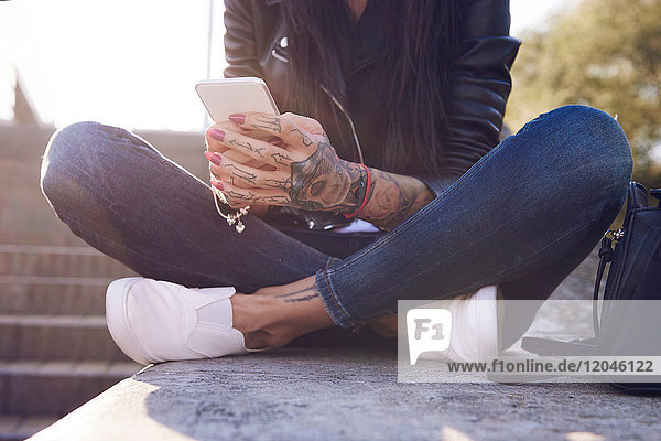 Junge Frau an der Wand sitzend,  Smartphone benutzend,  Tätowierungen auf der Hand,  niedriger Abschnitt